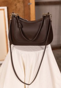 Bruine leren handtas met een gladde textuur, gebogen vorm, verstelbare riem en hardware in goudkleur. Logo gegraveerd aan de voorkant.