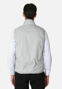 Gilet grigio chiaro, senza maniche, texture liscia, colletto alto, orlo elastico, indossato sopra una camicia bianca a maniche lunghe, design minimalista.