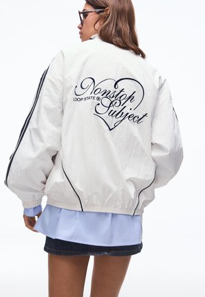 Žena nosí oversized bielu bundu s čiernymi pruhmi na rukávoch a nápisom „Nonstop Subject“ vo vnútri srdca na chrbte, doplnenú o rifľovú sukňu.