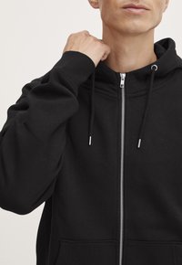Zwarte zip-up hoodie van zachte stof met een kangoeroezak, een capuchon met trekkoord en contrasterende zilveren ritsdetails.