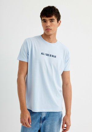 Camiseta de algodón de color azul claro con el texto "TODO LO QUE VEO ES AZUL" en azul oscuro, diseño de cuello redondo, mangas cortas, combinada con pantalones cortos de mezclilla claros.