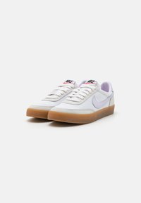 Adidași albi din plasă cu un swoosh Nike de culoare violet deschis și o talpă din cauciuc de culoare caramel. Prezintă șireturi albe și o etichetă de limbă branduită roșie și neagră.