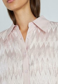 Lichtroze shirt met een glanzende stof en een zigzagpatroon, getextureerd met franjes, en afgewerkt met een klassieke kraag en knopen.