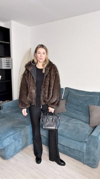 Καφέ μπουφάν από συνθετική γούνα με κουκούλα, μαύρο τοπ και παντελόνι, μαύρη τσάντα χειρός, όρθια σε ένα υφασμένο μπλε καναπέ σε έναν μίνιμαλ εσωτερικό χώρο.