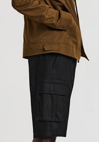 Braune Jacke mit Hand in der Tasche, getragen über schwarzen Cargo-Shorts mit Klappentasche an der Seite.