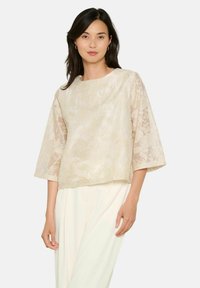 Top in pizzo floreale beige con maniche a tre quarti, scollo rotondo e tessuto testurizzato, abbinato a pantaloni bianchi. Nessun motivo visibile.