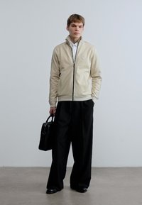 Μπεζ ζακέτα με φερμουάρ, με ριμπ μανσέτες και γιακά, μαύρο παντελόνι με φαρδύ πόδι και μαύρη δερμάτινη τσάντα. Λείο ύφασμα με ελάχιστες λεπτομέρειες.