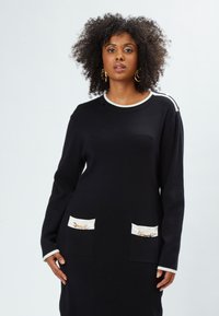 Abito nero in maglia con maniche lunghe, bordo bianco intorno al collo e tasche, con decorazioni in catena sui taschini.