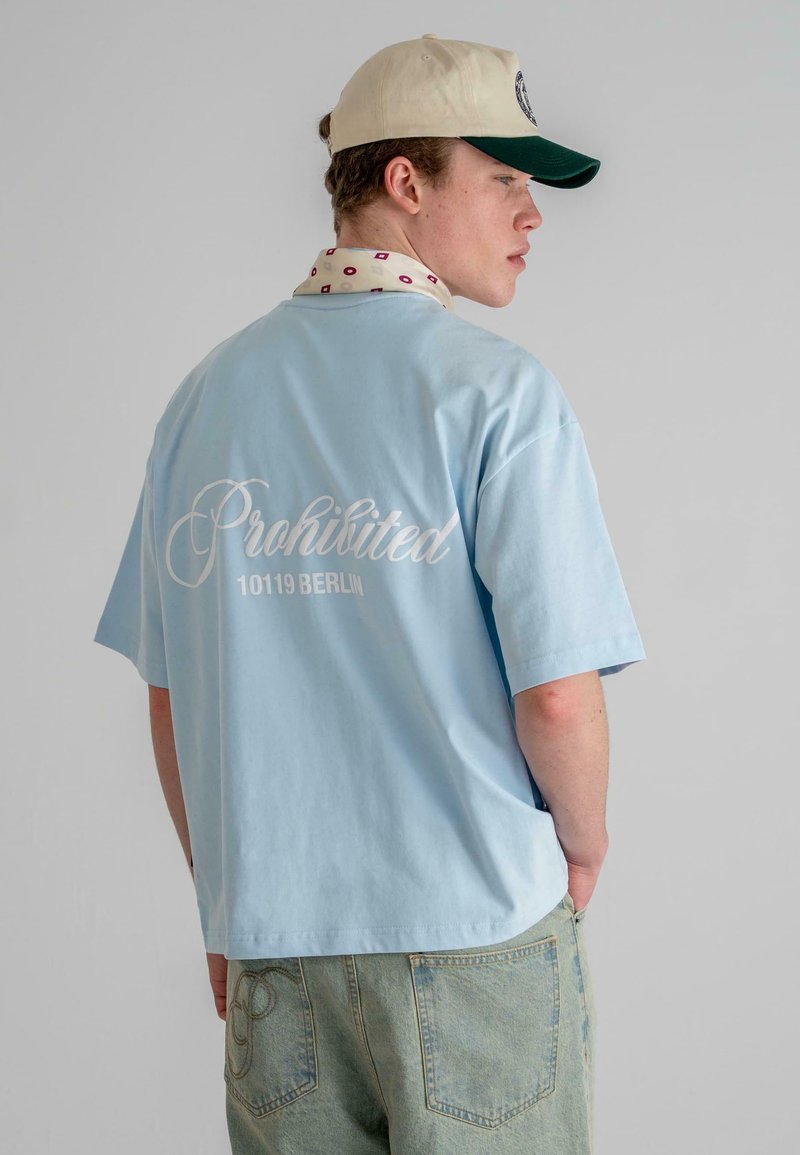 Light blue short-sleeve t-shirt featuring white script "Prohibited 10119 BERLIN" on the back, paired with green cap and patterned scarf.