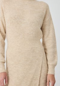 Maglione beige a maglia con collo alto, texture a coste e maniche lunghe, caratterizzato da un design drappeggiato e dettagli sulle cuciture laterali.