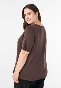 Pruun lühikeste varrukatega top lõdva istuvusega, ümar kaelus ja sile tekstuur, millel on kergelt kaardus ääreosa ja minimaalne õmblusdetail.