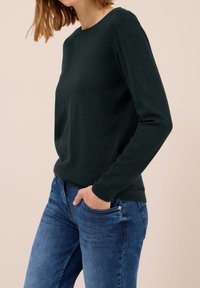 Femme portant un pull noir à manches longues et un jean bleu, une main dans la poche, sur un fond neutre.