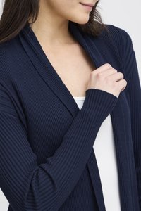 Tumesinine soonikkoes kardigan, pehme tekstuur, avatud esikujundus. Pikad varrukad tihedate mansettidega, kantud valge pluusi peal, esitades peenikesi detaile.