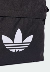 Black fabric backpack featuring a large white Adidas logo with three stripes beneath. The material has a textured finish.