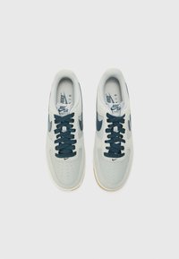 Nike Air Force 1 sneakers in light grey with dark blue accents, perforated toe box, thick laces, and rubber sole. Textured upper material.
