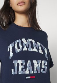 Námořnická modrá bavlněná tričko s velkým šedým a bílým grafickým nápisem "TOMMY JEANS" s texturovaným vzorem a malou vlajkovou aplikací na lemu.