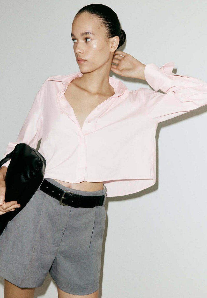Femme portant un chemisier court rose clair et un short gris avec une ceinture noire, tenant un sac noir et regardant sur le côté.