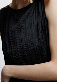 Vestido negro texturizado con pliegues verticales, escote redondo y diseño sin mangas. Tejido suave con un ligero brillo, que acentúa la forma.