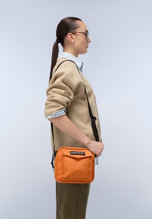 Kvinde med langt hår i hestehale iført solbriller, beige sweater over stribet skjorte, holder en orange crossbody taske med sort rem.
