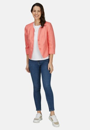 Lachende vrouw met lang donker haar, die een koraalblazer, wit shirt, blauwe jeans en witte sneakers draagt, staand tegen een witte achtergrond.