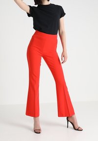 Červené upnuté zvonové kalhoty s vysokým pasem, hladkým povrchem a jednobarevným designem, v kombinaci s černým topem s krátkým rukávem a černými vysokými podpatky.