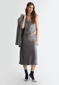 Gilet in denim con dettagli invecchiati, gonna midi grigia in satin, calze alla caviglia nere e scarpe con tacco alto argento, il tutto su uno sfondo neutro.