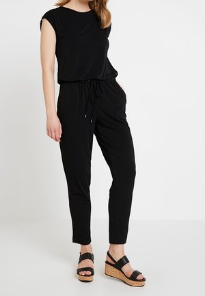 Kvinde iført en sort ærmeløs jumpsuit med snøre i taljen og sorte kilehælssandaler med korkplatform.