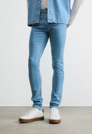 Hombre con jeans ajustados azul claro, zapatillas blancas con suelas marrones y chaqueta vaquera azul claro sobre una camiseta blanca, de pie sobre un suelo gris.