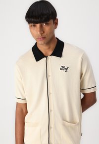 Kurzarm-Cardigan aus cremefarbener Wollmischung mit schwarzem Kragen und Besatz. Verfügt über Vordertaschen und ein gesticktes Logo auf der Brust.