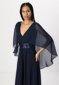 Navy gown with a deep V-neck and flowing sheer sleeves. Sequined waistband adds texture. Smooth, lightweight fabric. Elegant silhouette.