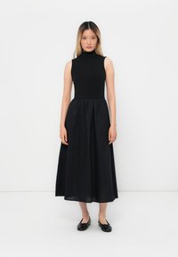 Nainen seisoo valkoista seinää vasten, päällään musta hihaton korkeakauluksinen mekko ja mustat ballerinat, katsoo eteenpäin neutraalilla ilmeellä.
