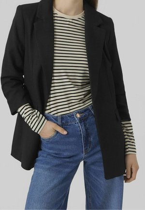 Personne portant un blazer noir sur une chemise beige et noire rayée à manches longues, avec un jean bleu taille haute, une main dans la poche.