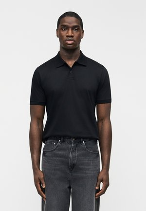 Noor mees seisab liikumatult, kandes musta lühikeste varrukatega polo särki ja pleekinud musta teksapükse, lihtsa heleda tausta ees.