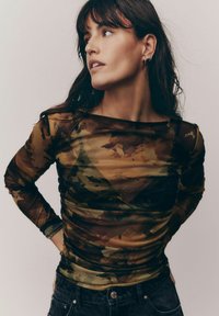 Halenka s dlouhým rukávem, průhledná, v maskáčovém vzoru se zemními tóny. Má nabíranou texturu, široký výstřih a slim fit, kombinovaná s tmavými džíny.