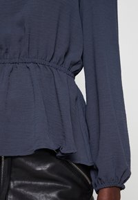 Blouse bleu foncé à manches longues avec taille élastique froncée et ourlet volanté portée sur un pantalon en cuir noir avec fermeture éclair visible.