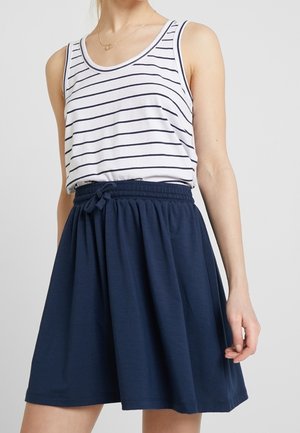 Kvinde iført en hvid tanktop med sorte horisontale striber, stoppet ned i et marineblåt skørt med snøre, foran en ensfarvet baggrund.