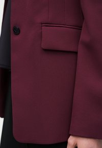 Κοντινό πλάνο στην τσέπη ενός μπλεζέρ βουργουνδίας με λεπτομέρεια καπακιού και μαύρο κουμπί, που δείχνει μέρος του μανικιού και του χεριού.