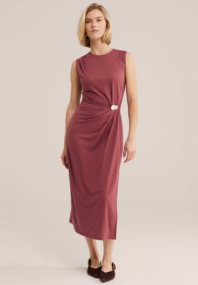 Femme portant une robe midi sans manches couleur vieux rose avec un détail twist sur le côté et une boucle circulaire, associée à des chaussures pointues marron foncé.