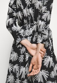 Černé šaty s bílým listovým vzorem, dlouhými rukávy a zvýrazněným pasem. Tkanina vypadá měkce s hladkým povrchem. Ruce složené za zády.