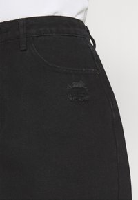 I jeans in denim nero presentano una toppa strappata sulla tasca anteriore. Il materiale è testurizzato con un taglio classico e dettagli di cucitura sottili.