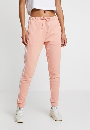 Kobieta w jasnoróżowych spodniach dresowych z białymi paskami po bokach i białych tenisówkach, krótki biały top, stoi prosto.