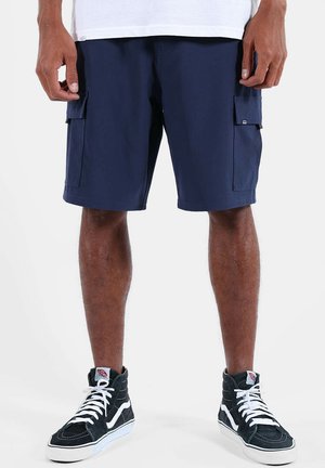 Homme portant un short cargo bleu marine et des baskets montantes noires à lacets blancs, debout devant un fond uni.