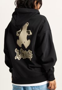 Schwarzer Hoodie aus dickem Stoff, mit einem großen beigen Grafik eines Bären und stilisiertem Text auf der Rückseite, der hintere Saum gerippt.