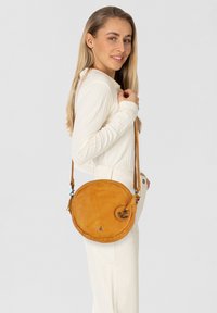 Borsa a tracolla rotonda in pelle gialla senape con superficie liscia, tracolla regolabile e hardware in metallo. Presenta un emblema decorativo a forma di ancora.