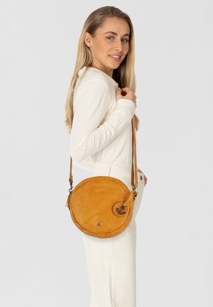 Borsa a tracolla rotonda in pelle gialla senape con superficie liscia, tracolla regolabile e hardware in metallo. Presenta un emblema decorativo a forma di ancora.