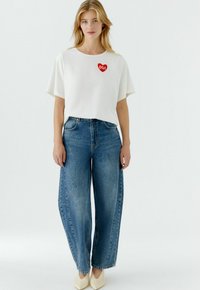 Witte T-shirt met korte mouwen en rode hartvormige "OUI" borduring, gecombineerd met hooggetailleerde, wijd uitlopende blauwe spijkerbroeken en schoenen in neutrale kleuren.