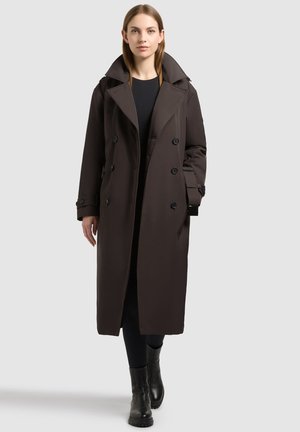 Femme aux cheveux raides portant un long manteau marron foncé à double boutonnage, des leggings noirs et des bottines noires sur un fond uni.
