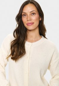 Femme aux longs cheveux bruns portant un cardigan crème à boutons, regardant légèrement sur le côté avec un fond clair neutre.