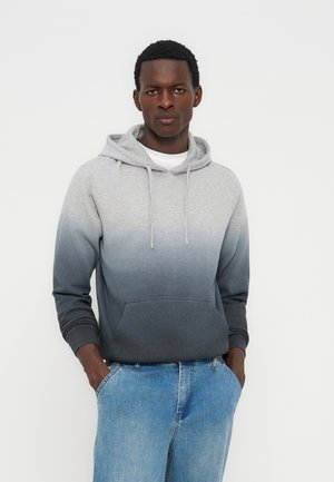 Mees, kes kannab hallist mustani ombréstiilset dressipluusi eest taskuga ja helesiniseid teksapükse, seisab käed taskutes valge tausta ees.