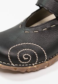 Zwarte leren schoen met uitsnijdingen, voorzien van beige spiraalstiksels en een gestructureerde bruine zool. Klittenbandsluiting voor een veilige pasvorm.